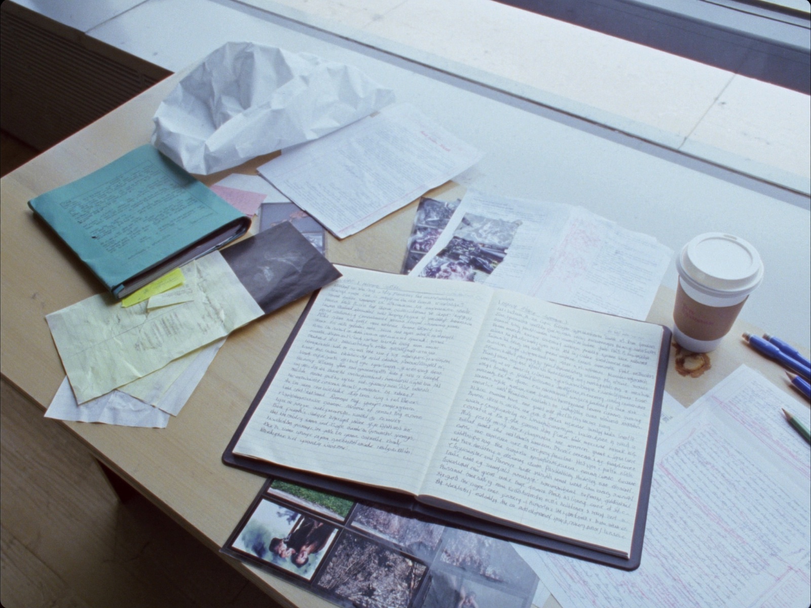 A documentary producer's cluttered prep desk by a window: an open handwritten notebook, a treatment folder, fanned reference photos, sticky notes, a crumpled page, pencils, and a coffee cup
